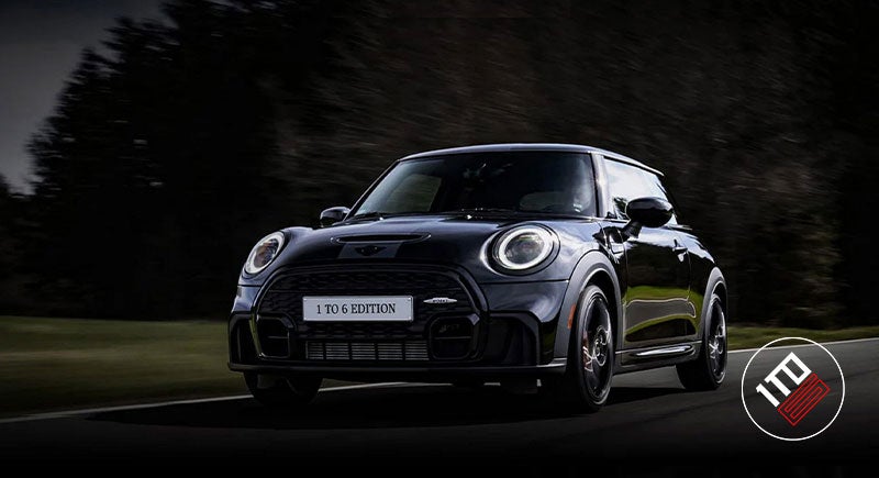 Front 3/4 angle view of JCW 1to6 driving down a country road with MINI John Cooper Works badge in bottom right, including 'JOHN COOPER' red text positioned horizontally on top of 'WORKS' in black text along with red lines extending off either side.